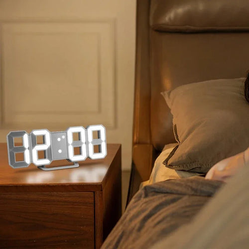 White 3D LED digital alarm clock displaying 12:00 on a wooden bedside nightstand table next to a bed with pillows under warm ambient bedroom lighting.