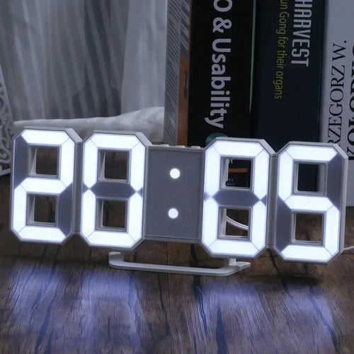 White 3D LED digital alarm clock displaying 12:00 on a wooden bedside nightstand table next to a bed with pillows under warm ambient bedroom lighting.